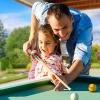 Father leaning over his daughter at the billiards table helping her to make a shot.
